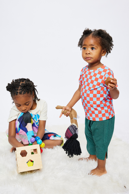 18 month old girl with black curly hair holding a muffin in one hand while her 5 year old sister is playing next to her. she is wearing a red and blue checker tshirt made from organic cotton.