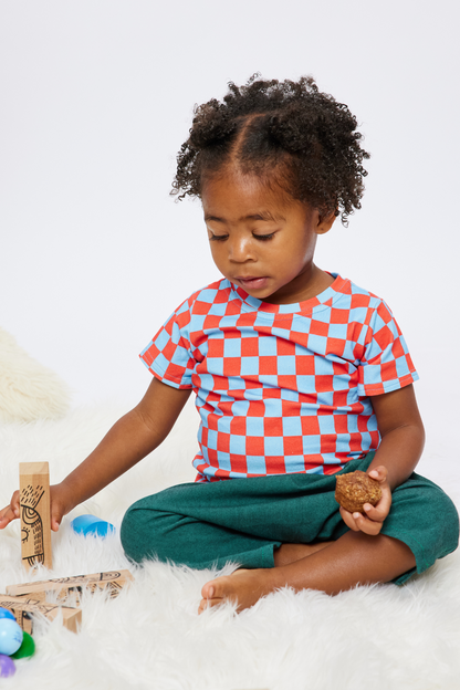 18 month old girl with black curly hair holding a muffin in one hand and a wooden block in the other. she is wearing a red and blue checker tshirt made from organic cotton and cotton/bamboo pants.