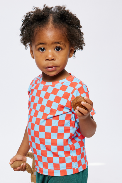 18 month old girl with black curly hair holding a muffin in one hand and looking at the camera. she is wearing a red and blue checker tshirt made from organic cotton.
