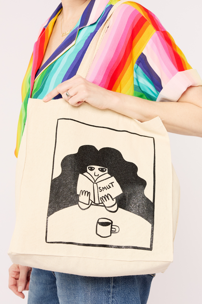 cream cotton tote bag with an illustration of a woman with dark curly hair reading a book titled smut