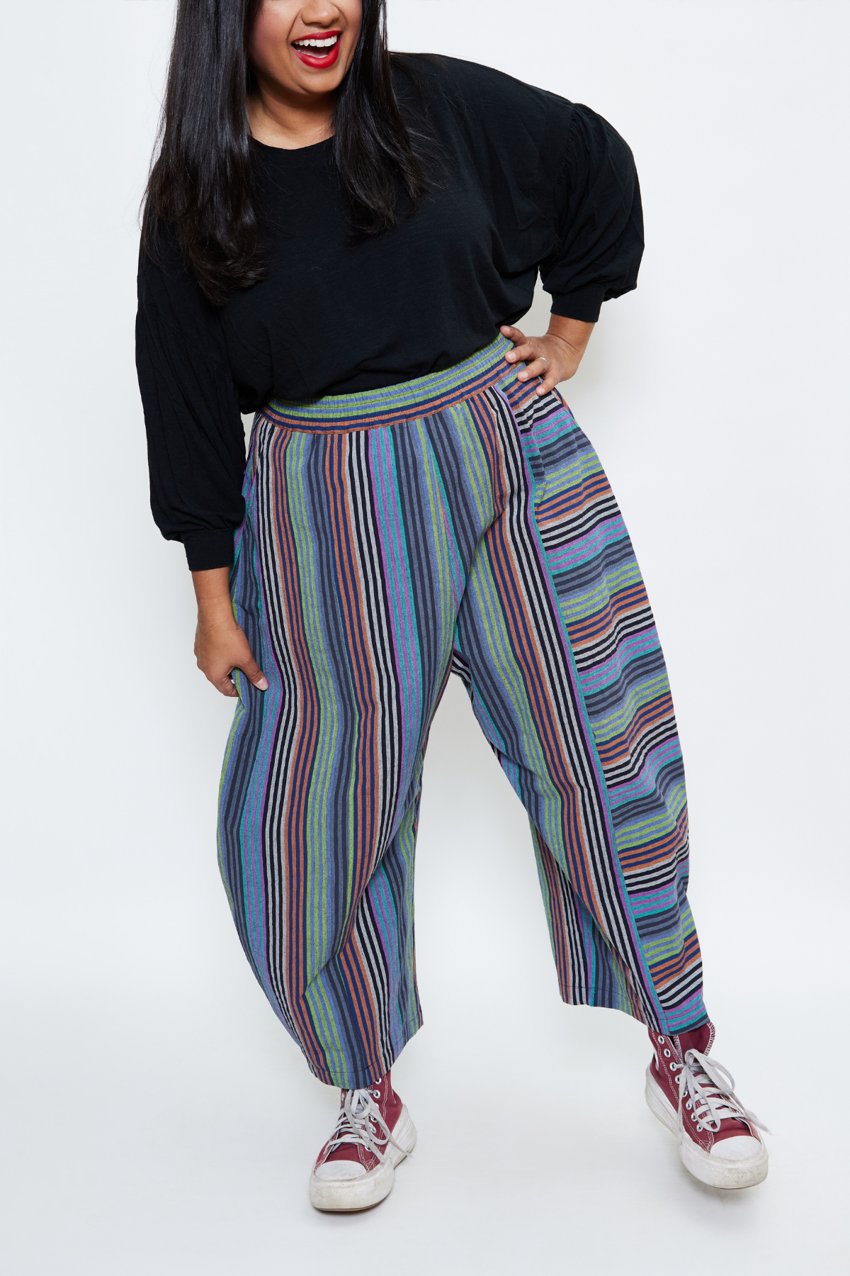 fun woman standing and leaning on one leg while wearing barrel leg sustainable pants, a black tee, and cool sneakers. she's laughing.