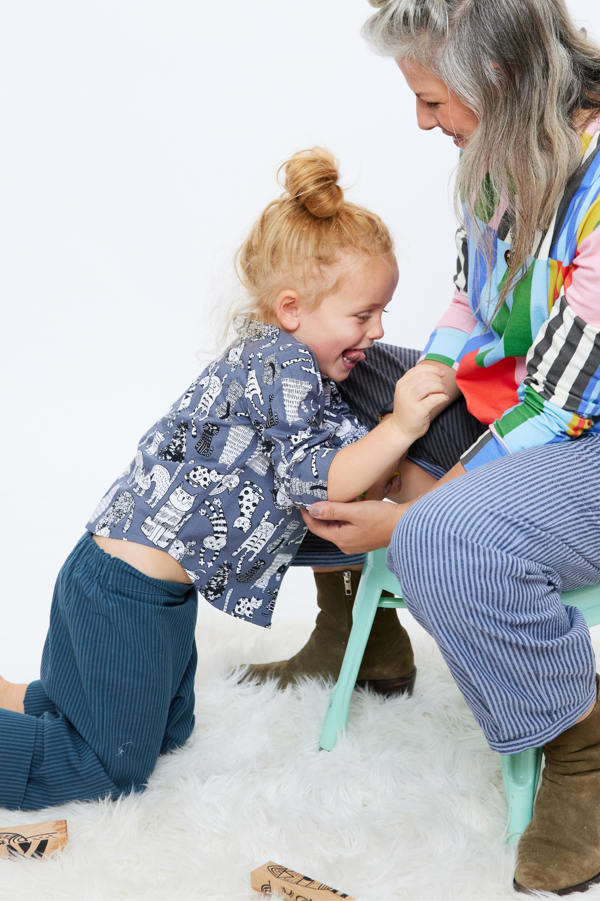 four year old child with long hair up in a bun is wearing a gray collar-less button down shirt with a chest pocket and cuffs rolled up. There are illustrated cats on the shirt. The child is sitting and playing with his mom on the floor. 