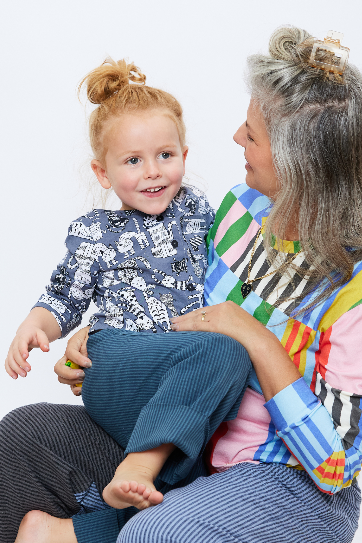 four year old child with long hair up in a bun is wearing a gray collar-less button down shirt with a chest pocket and cuffs rolled up. There are illustrated cats on the shirt. He is sitting on his mom's lap. She is wearing a brightly colored long sleeve t-shirt.