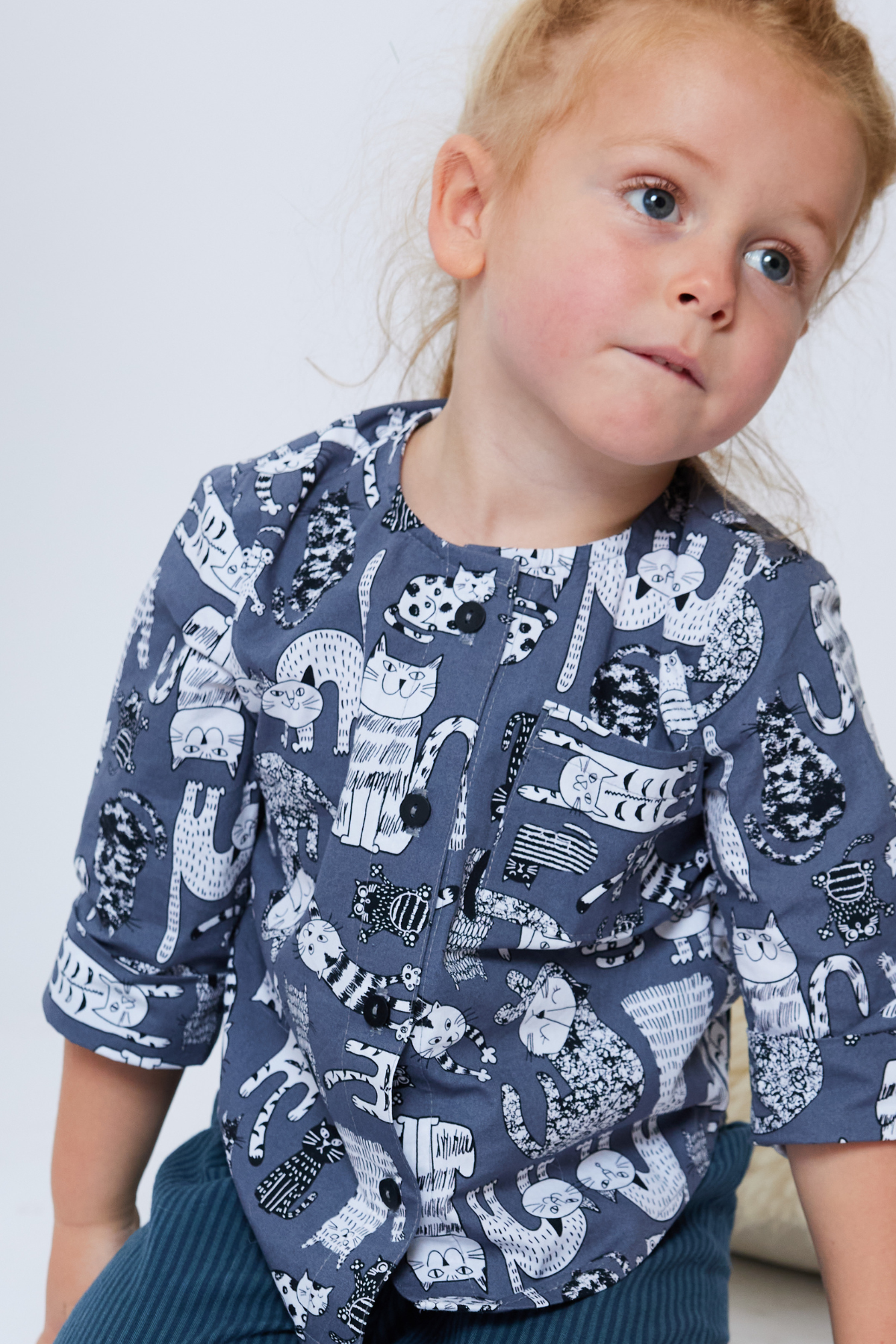 four year old child with long hair up in a bun is wearing a gray collar-less button down shirt with a chest pocket and cuffs rolled up. There are illustrated cats on the shirt.