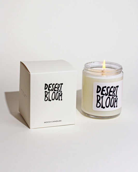 A clear glass jar soy candle with a label that reads 'DESERTBLOOM', placed next to its white box packaging 