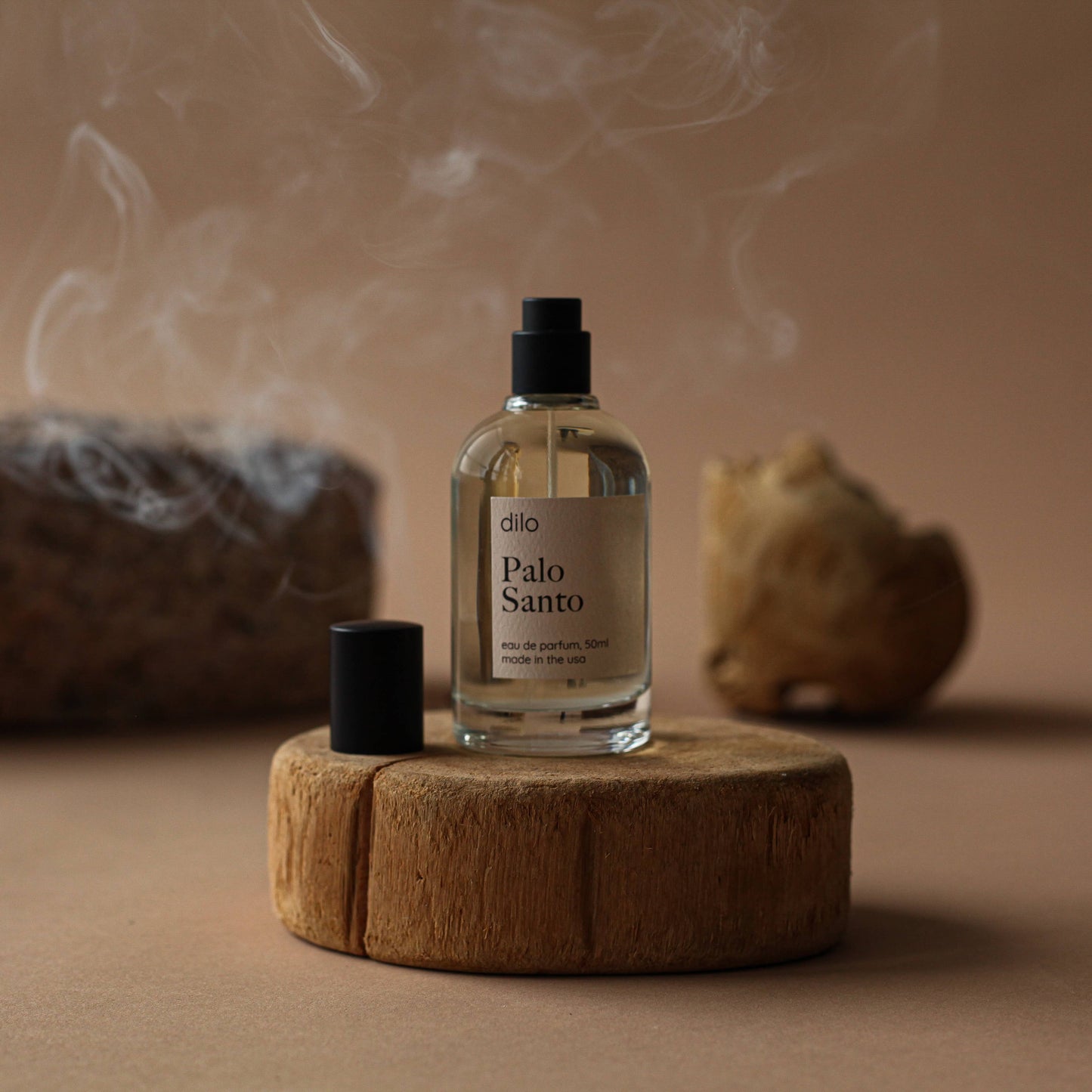 A clear glass bottle of 'Palo Santo' eau de parfum on a wooden platform, with a matte black sprayer and cap, surrounded by a warm-toned background.