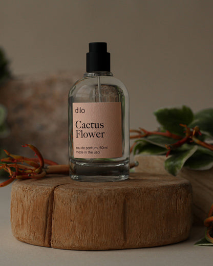 A 50ml clear glass vessel of 'Cactus Flower' unisex eau de parfum with a matte black fine mist sprayer and cap, displayed on a wooden stand with green foliage in the background.